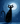 A stylized illustration depicts a black cat sitting upright, facing forward, with wide white eyes and prominent whiskers. It is silhouetted against a deep blue, textured night sky filled with scattered white stars, with a large, bright full moon positioned behind its head to the upper right. The cat's dark body also features small scattered white and light blue stars, and its long, curved tail rests gracefully beside it, with a subtle shadow beneath its paws.
