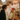 A young man and woman are laughing together in a warmly lit kitchen decorated for Christmas. The man, with curly dark hair, wears a dark sweater, while the blonde woman wears a cream ribbed sweater and a festive red knit hat with colorful patterns, both looking at each other with wide smiles. Behind them, a white refrigerator is covered with magnets, and shelves are adorned with numerous string lights and green garlands, creating a cozy and joyful holiday scene.