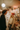 A young man and woman are laughing together in a warmly lit kitchen decorated for Christmas. The man, with curly dark hair, wears a dark sweater, while the blonde woman wears a cream ribbed sweater and a festive red knit hat with colorful patterns, both looking at each other with wide smiles. Behind them, a white refrigerator is covered with magnets, and shelves are adorned with numerous string lights and green garlands, creating a cozy and joyful holiday scene.