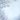 A beautiful winter scene featuring delicate tree branches covered in glistening white frost. The branches frame the top and bottom-left of the image, while the center and right reveal a soft, light blue background with gentle, sparse snowflakes falling. This serene image evokes a peaceful and cold winter day.
