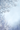 A beautiful winter scene featuring delicate tree branches covered in glistening white frost. The branches frame the top and bottom-left of the image, while the center and right reveal a soft, light blue background with gentle, sparse snowflakes falling. This serene image evokes a peaceful and cold winter day.