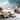 A close-up photograph displays a collection of beautifully decorated Christmas cookies on a light grey wooden surface. The prominent cookie in the foreground is a light blue snowflake, intricately decorated with white icing and sparkling gold sprinkles. In the soft-focus background, warm golden string lights create a magical bokeh effect, complemented by rustic pinecones dusted with white.