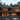 A two-story modern house with a minimalist design, featuring dark gray concrete and natural wood paneling, stands amidst a dense pine forest at dusk. Large floor-to-ceiling windows reveal warm, illuminated interiors, including living and dining areas, while a small deck extends onto a sandy, natural ground. The scene evokes a sense of luxurious tranquility and contemporary living in nature.