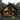 A striking modern dark-colored A-frame cabin is nestled in a dense green pine forest, captured at dusk. The large, multi-pane windows reveal a warm, glowing wooden interior, creating a cozy contrast with the dark exterior and the muted twilight sky. A spacious wooden deck with dark outdoor furniture extends from the house into a natural foreground of green grass and scattered large rocks.