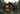 A striking modern dark-colored A-frame cabin is nestled in a dense green pine forest, captured at dusk. The large, multi-pane windows reveal a warm, glowing wooden interior, creating a cozy contrast with the dark exterior and the muted twilight sky. A spacious wooden deck with dark outdoor furniture extends from the house into a natural foreground of green grass and scattered large rocks.