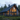 A modern A-frame cabin stands nestled in a dense evergreen forest at dusk. The house features dark vertical wood siding, a dark corrugated metal roof with two chimneys, and large, triangular floor-to-ceiling windows glowing with warm, golden interior light. A wooden deck with contemporary outdoor furniture and a glass railing extends in front of the house, surrounded by lush green foliage and tall pine trees under a soft, cloudy sky.