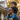 A smiling Black father with glasses carries his young Black son on his shoulders, both looking happily towards the right. The son wears a black shirt, blue pants, and vibrant multi-colored sneakers, while the father wears a dark grey shirt. Golden hour sunlight backlights them against a soft, blurred background of a house and autumn foliage, conveying a warm and joyful family moment.