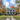 A classic white two-story house with a green metal roof and black shutters stands centered in an autumn landscape. A dark brown dog sits on a gravel driveway leading to the front porch, which has white columns and red Adirondack chairs. The house is surrounded by vibrant orange, yellow, and red fall foliage, green grass with scattered leaves, and a bright blue sky with white clouds.