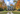 A beautiful white colonial-style house with a green metal roof and black shutters stands prominently at the end of a symmetrical gravel driveway. A dark brown dog sits facing the house on the driveway. The entire scene is framed by a dense forest of trees bursting with vibrant orange, yellow, and green autumn leaves under a bright blue sky dotted with white clouds.
