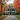 A stately, symmetrical white colonial-style house with a green roof and black shutters stands centered at the end of a long, gravel driveway. Vibrant red, orange, and yellow autumn trees frame the scene, with fallen leaves scattered on the green grass and textured driveway. A small, golden-colored dog is sitting calmly on the driveway near the house's entrance.