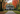 A stately, symmetrical white colonial-style house with a green roof and black shutters stands centered at the end of a long, gravel driveway. Vibrant red, orange, and yellow autumn trees frame the scene, with fallen leaves scattered on the green grass and textured driveway. A small, golden-colored dog is sitting calmly on the driveway near the house's entrance.