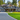 A large, modern two-story luxury home is centered in the frame, featuring dark gray shingle siding, white trim, and natural wood accents. A curving aggregate driveway leads to a two-car garage on the left, while a meticulously manicured green lawn stretches across the foreground. The house is beautifully framed by vibrant autumn foliage in shades of orange, yellow, and red, and tall evergreen trees under a bright, clear sky, creating a serene and luxurious setting.