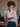 A smiling young woman with curly brown hair, wearing a blush pink blazer with gold embroidery, a white shirt, and navy trousers, looks down at a dark smartphone in her right hand. Her genuine smile reveals dimples, and she wears a gold necklace and earring. The background is a softly blurred, warm-toned modern office setting.