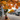 A wide shot of a charming small town street during autumn, adorned with a canopy of vibrant orange and yellow leaves overhead and scattered on the ground. Historic multi-story buildings with varied facades, including brick, clapboard, and shingle, line both sides of the street, with numerous cars parked along the curb. An American flag flies from a pole attached to a building on the left, and a planter with bright pink flowers adds a pop of color in the foreground on the sidewalk.