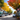 A wide shot of a charming small-town main street bustling with vibrant autumn colors under a clear blue sky. On both sides, historic buildings with storefronts and colorful awnings line the street, while large trees display brilliant golden yellow, orange, and red fall foliage. An American flag hangs from a tree on the right, and flower boxes with autumn blooms adorn the sidewalks, with several cars parked along the road receding into the distance.