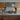 A happy man and woman are at a long white kitchen island in a brightly lit modern kitchen. A dark laptop sits between them; the woman types while the man, wearing a denim shirt, smiles at her. The kitchen features sleek light wood cabinets, stainless steel appliances, and a minimalist design, creating an inviting and productive home atmosphere.
