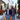 A diverse couple, a man and a woman, walk smiling down a sunlit, upscale urban shopping street, carrying multiple brightly colored shopping bags. The woman, on the left, has curly brown hair and wears a blue puffer jacket with a grey scarf, while the man on the right wears a similar jacket over a reddish-brown shirt and blue jeans. They are framed by elegant white buildings with large display windows, lush green trees, and vibrant red flowers in white planters along the sidewalk.