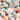 A top-down, close-up view shows a fluffy white bunny's head and pink ears emerging from a vibrant pile of colorful pastel Easter eggs. The eggs are light blue, pale yellow, peach, and white with multi-colored speckles, interspersed with small white spring flowers, creating a whimsical and festive scene.