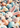 A top-down, close-up view shows a fluffy white bunny's head and pink ears emerging from a vibrant pile of colorful pastel Easter eggs. The eggs are light blue, pale yellow, peach, and white with multi-colored speckles, interspersed with small white spring flowers, creating a whimsical and festive scene.