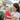 A young woman with dark hair, wearing a white t-shirt, holds a fair-haired toddler in a red t-shirt. The woman smiles looking down at the child, while the toddler grins broadly looking directly at the viewer. They are outdoors in natural sunlight, with a softly blurred background of a light-colored house and green foliage.