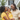 A Black mother, father, and young daughter are captured in a bright, outdoor portrait. The mother, with dreadlocks and wearing a yellow top, holds her smiling daughter, who is in a ruffled yellow dress and looks directly at the viewer. The father, also with dreadlocks and wearing a beige shirt, smiles warmly down at his daughter, with a modern white and black house, green lawn, and palm trees visible in the blurred background, suggesting a serene home environment.