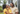 A Black mother, father, and young daughter are captured in a bright, outdoor portrait. The mother, with dreadlocks and wearing a yellow top, holds her smiling daughter, who is in a ruffled yellow dress and looks directly at the viewer. The father, also with dreadlocks and wearing a beige shirt, smiles warmly down at his daughter, with a modern white and black house, green lawn, and palm trees visible in the blurred background, suggesting a serene home environment.