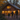 A family of four, including two adults and two young children, walks hand-in-hand on a gravel path towards a rustic wooden cabin at dusk. The cabin's windows and exterior lights glow warmly, contrasting with the dark forest surrounding it, which features trees with golden autumn leaves.