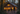 A family of four, including two adults and two young children, walks hand-in-hand on a gravel path towards a rustic wooden cabin at dusk. The cabin's windows and exterior lights glow warmly, contrasting with the dark forest surrounding it, which features trees with golden autumn leaves.