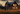 A family of four, consisting of two adults and two young children, walks away from the viewer through a vibrant golden field of tall grass towards a cozy, dark wooden house. The house's windows glow warmly with interior light, contrasting with the deep blues and oranges of the twilight sky, while the family's silhouettes are softly illuminated by the last rays of the setting sun, evoking a profound sense of peace and belonging at home.