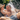 A close-up photo captures a father and his young daughter sharing a moment of pure joy outdoors on a sunny day. Both are laughing with mouths wide open as water splashes over them, with visible droplets on their wet skin and clothes. The girl wears a straw hat and a white top, hugging her father who is bare-chested with denim shorts, set against a blurred green foliage background.