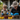 An elderly woman and a young boy, both wearing bright blue gloves, are deeply engrossed in potting plants on a light-colored wooden table outdoors. The woman, with a straw hat and glasses, is seen leaning over, actively involved in the task while the young boy, in beige overalls, mimics her actions, scooping soil. Various colorful pots containing small green seedlings or soil are arranged on the table, highlighting a shared, nurturing gardening activity between generations.