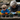 A senior woman with gray hair and glasses, wearing a dark jacket and light blue gloves, is shown leaning over a wooden table, carefully planting a small seedling. Next to her, a young boy in a blue jacket and bright orange gloves is also focused on scooping soil into a small pot. Several potted plants, some with yellow flowers, are visible on the table, indicating a shared gardening activity outdoors, with a blurred stone building and greenery in the background.