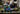 A senior woman with gray hair and glasses, wearing a dark jacket and light blue gloves, is shown leaning over a wooden table, carefully planting a small seedling. Next to her, a young boy in a blue jacket and bright orange gloves is also focused on scooping soil into a small pot. Several potted plants, some with yellow flowers, are visible on the table, indicating a shared gardening activity outdoors, with a blurred stone building and greenery in the background.