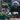 A close-up shot of an older woman with grey hair and a young child with brown hair, both wearing light-colored gloves and focused on potting plants on a weathered wooden table outdoors. The woman, on the right, uses a small trowel to plant a seedling into a small terracotta pot, while the child, on the left, carefully tends to a green plant pot filled with green foliage and delicate yellow flowers. The background is a softly blurred garden with various plants and a fence.