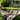 A comfortable cream-colored string hammock hangs in a vibrant green backyard, generously filled with multiple colorful, patterned quilts and an assortment of decorative throw pillows. A small wooden side table sits nearby, displaying two clear glasses of orange juice and a wooden bowl brimming with fresh red cherries. In the softly blurred background, a traditional brick house with white columns is visible through verdant trees, bathed in natural light.