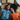 An overhead shot of a happy young adult Black couple lying on the floor, surrounded by cardboard moving boxes and soft pillows. The man, wearing a blue denim shirt, smiles widely, while the woman, in an orange ribbed shirt, rests her head on his shoulder, also smiling brightly at the camera. The scene exudes joy and excitement about starting a new chapter together.