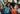 An overhead shot of a happy young adult Black couple lying on the floor, surrounded by cardboard moving boxes and soft pillows. The man, wearing a blue denim shirt, smiles widely, while the woman, in an orange ribbed shirt, rests her head on his shoulder, also smiling brightly at the camera. The scene exudes joy and excitement about starting a new chapter together.