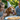 A brown and white spaniel dog wears a festive white party hat with colorful polka dots and silver tinsel trim, its tongue playfully out. The dog is looking slightly upwards, surrounded by smiling people; a young girl with blonde hair laughs on the left, and a woman with long brown hair smiles gently on the right, both looking fondly at the dog. The joyful outdoor scene is bathed in natural light, with a blurred green foliage background and a blurry white plate with food in the foreground.