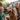A happy Golden Retriever dog wearing a vibrant, patterned party hat with a red pom-pom is surrounded by four smiling children in an outdoor setting. The dog has its mouth open and tongue slightly out, appearing content, while the children gaze at it with joyful expressions. The scene is illuminated by warm natural light, conveying a heartwarming sense of celebration and companionship between pets and kids.
