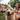 A golden retriever dog, wearing a colorful party hat with an orange pom-pom, looks directly forward with its tongue sticking out. In the blurred background, a smiling woman and two young children are visible, enjoying an outdoor celebration on a sunny day with green grass and trees.