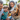 A close-up, eye-level shot of a happy white and reddish-brown dog wearing a colorful party hat with a sparkly dark blue pom-pom, its mouth slightly open and tongue visible in a contented expression. In the blurred background, a smiling man, woman, and two children are visible, looking fondly at the dog. The scene is set outdoors with soft green foliage, conveying a joyful family pet celebration.