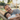 A man with a beard, wearing a grey t-shirt, and a young boy in a white striped t-shirt are resting together in a beige woven hammock. Both have their eyes closed and are smiling gently, appearing very content. The scene is outdoors, with warm, golden sunlight shining from behind them, creating a soft glow, and a blurred green garden visible in the background.