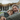 A close-up horizontal shot shows a smiling adult man with gray hair and beard embracing a young boy with blonde hair and freckles, both with their eyes closed in contentment. They are resting together in a beige woven rope hammock. The man wears a textured olive green sweater, and the boy wears a dark green puffer jacket, with a blurred modern house and green lawn in the background.