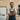 A smiling man with a beard, glasses, and messy brown hair stands with his arms crossed in a bright, modern kitchen. He is wearing blue denim overalls with yellow buckles over a white shirt. The background features light wooden cabinetry, a white refrigerator, built-in ovens, and a countertop with kitchen tools, suggesting a skilled individual in a contemporary home or workspace.