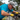 A close-up image shows a man with a beard and glasses, wearing a blue shirt and yellow work gloves, diligently cleaning a black rain gutter on a house roof. His hands are covered in grime, and foamy water splashes within the dirty gutter. The background features a brown shingle roof, light-colored house siding, and blurred green trees under an overcast sky.
