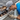 A close-up shot features a man in a blue shirt, grey baseball cap, and dirty work gloves intently leaning over a clogged residential gutter. The gutter is filled to the brim with wet, decomposing brown leaves and dark debris, with water visible among the organic matter. The grey asphalt shingles of the roof are visible in the background, emphasizing the outdoor home maintenance setting.