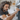 A man with dark, curly hair and a beard, wearing a dark gray t-shirt, intently looks down as he interacts with a silver front-loading washing machine. His right hand is near the open door of the machine, while his left hand holds a clear plastic bag near the opening, suggesting he is loading laundry. The background shows a softly blurred home interior with shelves and natural light.