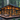 A striking modern cabin with extensive glass windows and warm wooden cladding is situated in a dense pine forest. The interior glows with a soft, inviting orange light, while a large wooden deck in the foreground features contemporary outdoor furniture including sofas, lounge chairs, and a dining set, surrounded by tall trees and scattered rocks, evoking a sense of peaceful luxury.