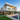 A stunning, multi-story modern luxury home with a white exterior and extensive floor-to-ceiling glass windows, situated on a hillside. In the foreground, an infinity pool with a concrete waterfall feature and a neatly manicured lawn with stepping stones are visible, bordered by lush green landscaping. The interior spaces are warmly illuminated, and a sprawling city skyline stretches into the distance under a gradient twilight sky.