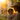 A warmly lit close-up of a smiling mother in a straw hat holding her baby outdoors during golden hour. The intense sunlight backlights them, creating a radiant halo effect around their hair and shoulders, with shimmering water droplets visible against lush green foliage in the left background. The baby, dressed in light fabric, looks towards the viewer with wide eyes while the mother, wearing a floral-patterned dress, gazes lovingly at the child.